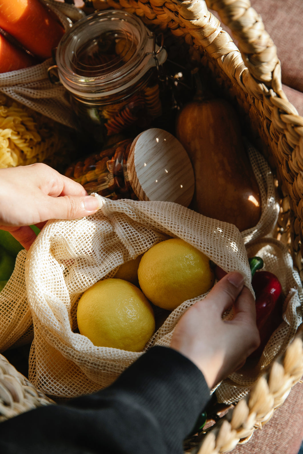 Cotton produce bags with fruit inside, within a basket of various jars and market items, with hands reaching inside the basket